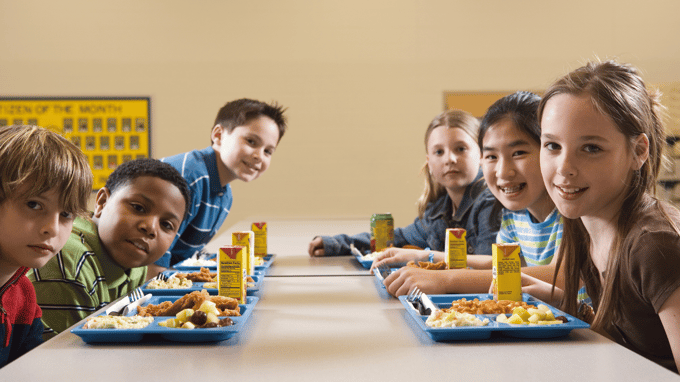 6 children, 3 on one side of table and 3 on other side. Each has a blue tray with food in front of them.