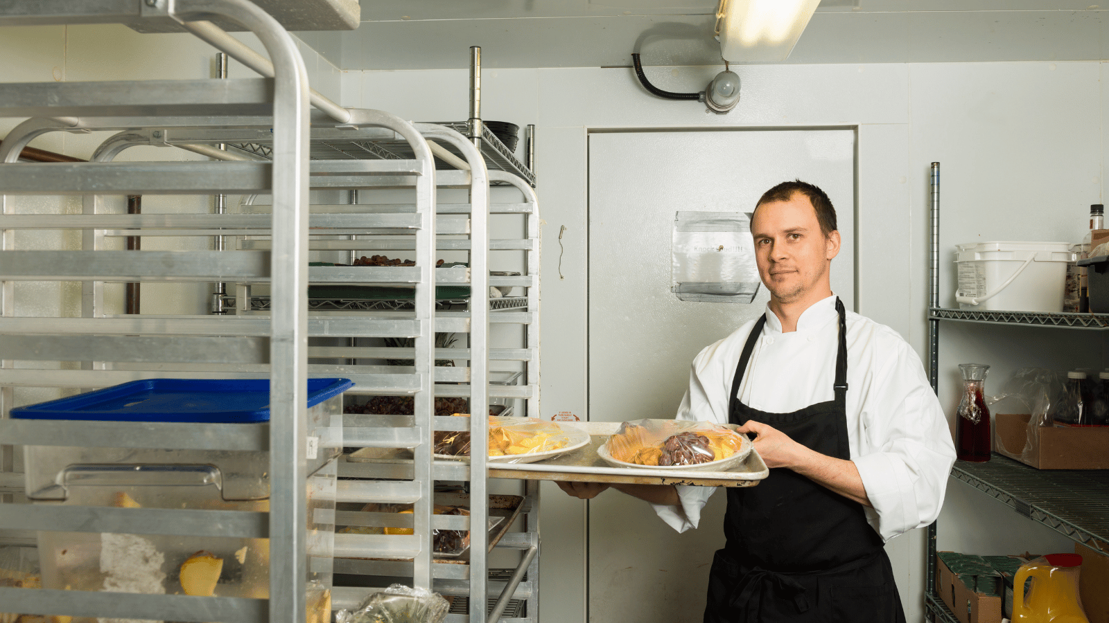 Worker in white shirt and black apron holding a tray inside a walk-in cooler. On either side are rolling racks and shelving.