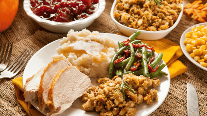 Close up of white plate with sliced turkey, mashed potatoes with gravy, stuffing, and green beans. Two forks are to the left of the plate, knife is to the right of the plate and the plate sits on a yellow cloth napkin. Above the plate in small white containers is cranberry sauce, stuffing, and corn.