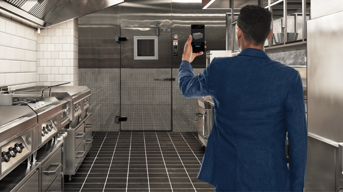 Man with blue suit jacket has back to viewer. He holds us a cell phone towards a walk-in cooler in a commercial kitchen.