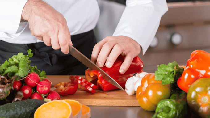 Hands with white chef's coat cutting red pepper on wood cutting board. Other veggies surround it.