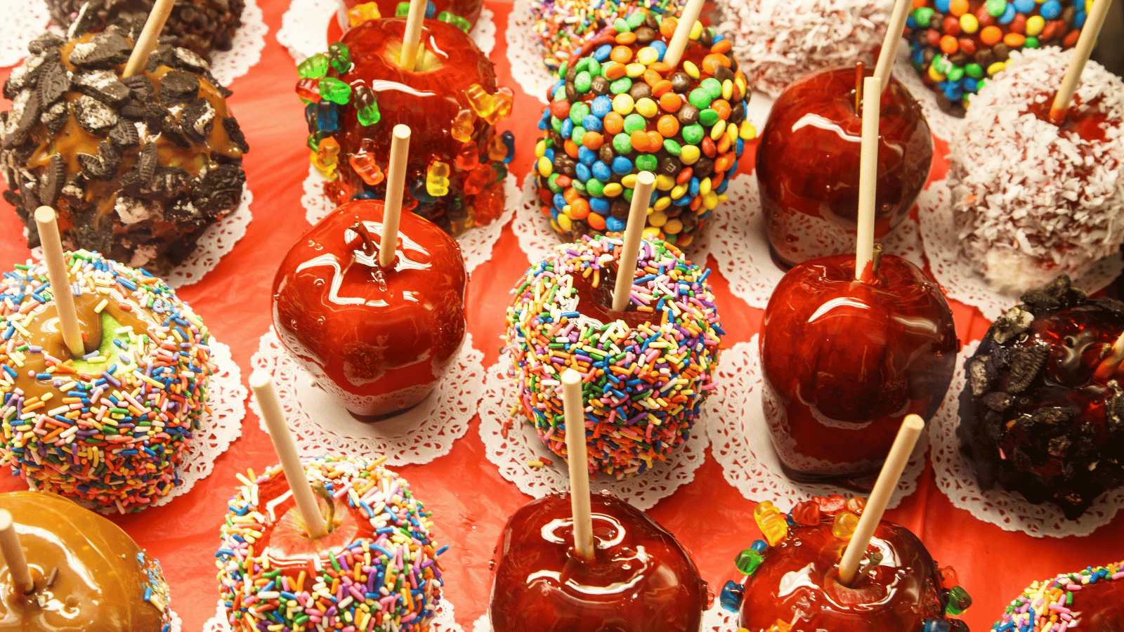 Multiple candy and caramel apples on white doilies. Some apples also have add-ons like gummy bears, crushed Oreos, and sprinkles.